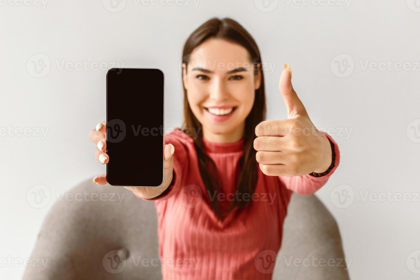 Happy Caucasian lady showing cellphone with empty screen and gesturing thumb up, recommending new mobile app or website on white studio background, mockup for advertisement photo