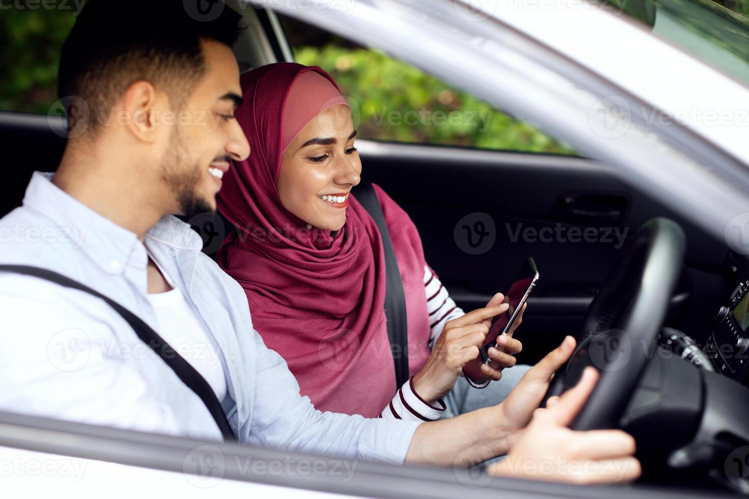 Young Muslim Spouses Riding Car And Using Smartphone Together, Happy Middle Eastern Couple Browsing Online Maps Or Gps Navigation On Mobile Phone, Planning Travel Route, Through Window Shot photo