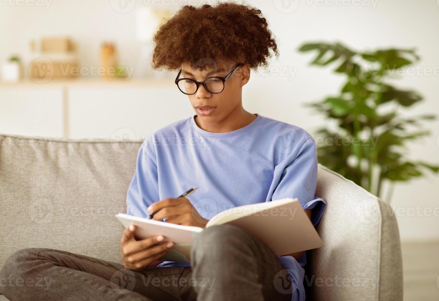Focused black teenager in glasses studying with notebook, writing down data, preparing for exam indoors. African American youth making homework, studying alone during covid lockdown photo