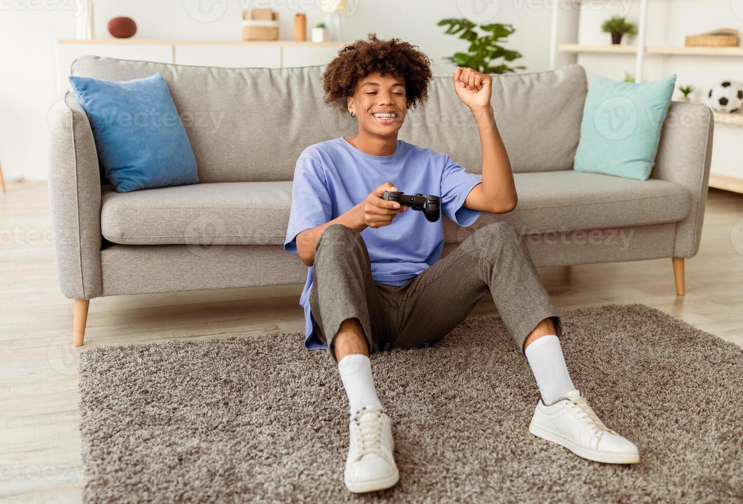 Full length of cheery black adolescent enjoying videogame on playstation, having fun at home. Positive Afro teenager with joystick sitting on floor, playing computer arcade. Lockdown hobbies concept photo