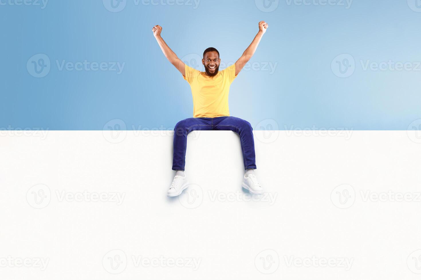 Advertising billboard template. Young African American guy sitting on top of blank white poster with mockup, lifting hands up in excitement on blue studio background, space for design photo