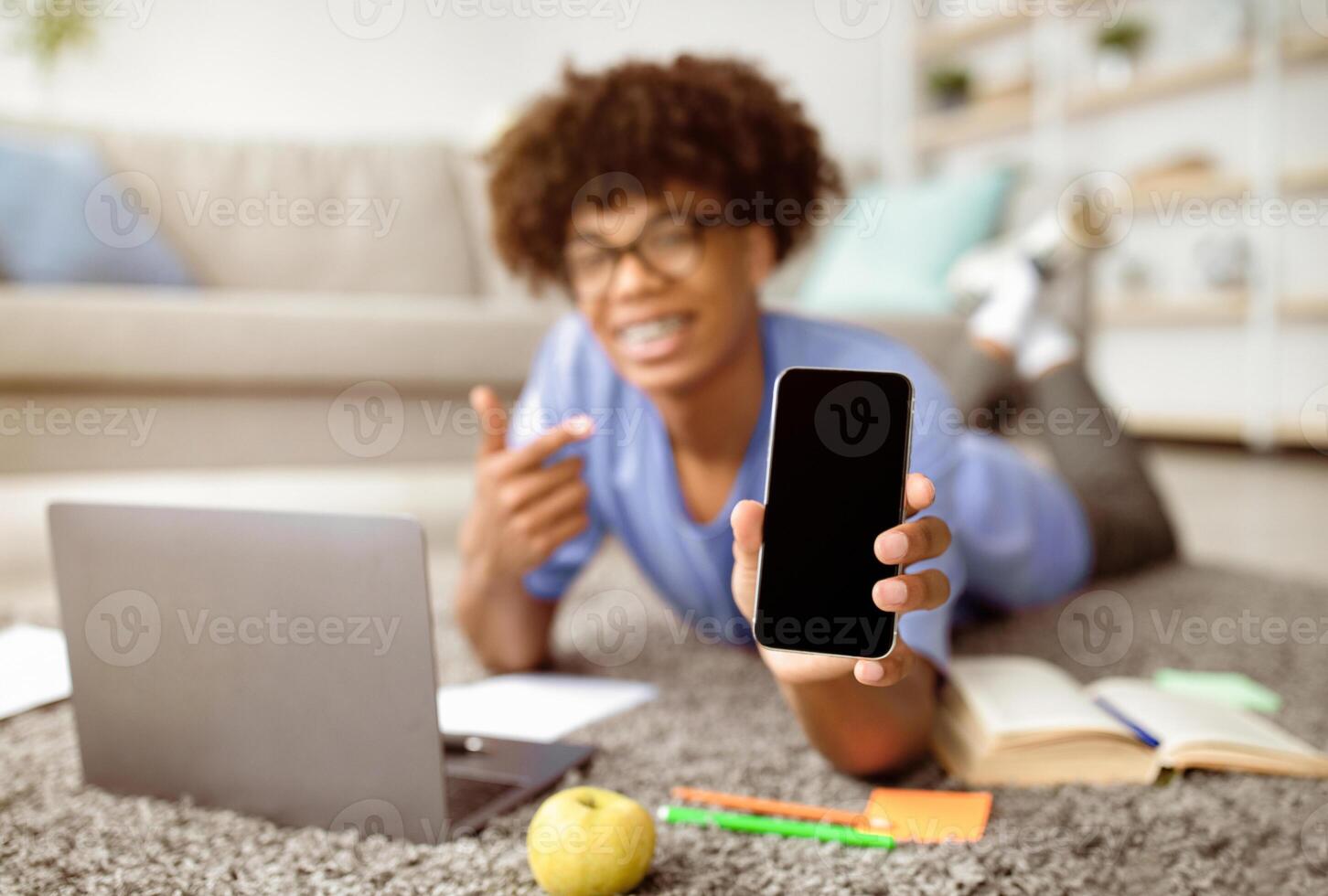 Positive black teenager lying on floor with laptop, pointing at smartphone with blank screen at home, mockup for mobile app or website design. Remote studies during coronavirus lockdown concept photo