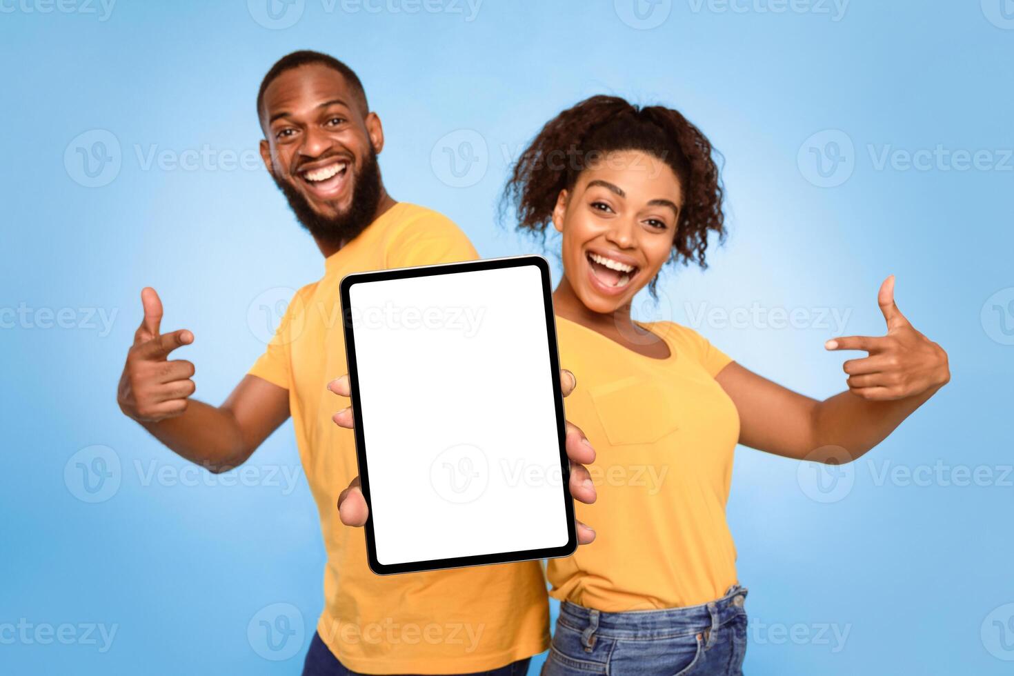 Joyful African American couple pointing at tablet screen with mockup for design, advertising new app or website over blue studio background. Touch pad display template, space for your ad photo