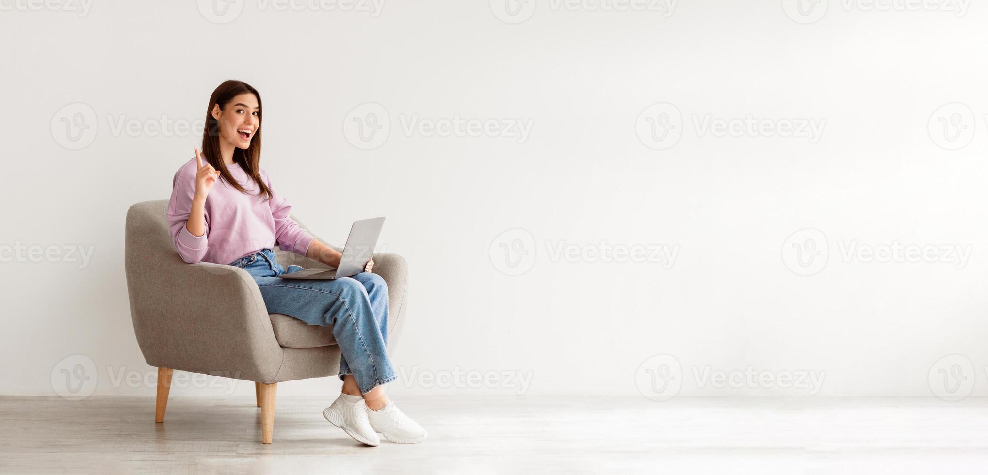 Millennial Caucasian woman having creative idea, gesturing eureka, sitting in armchair with laptop pc against white studio wall, copy space. Young lady finding solution to problem photo