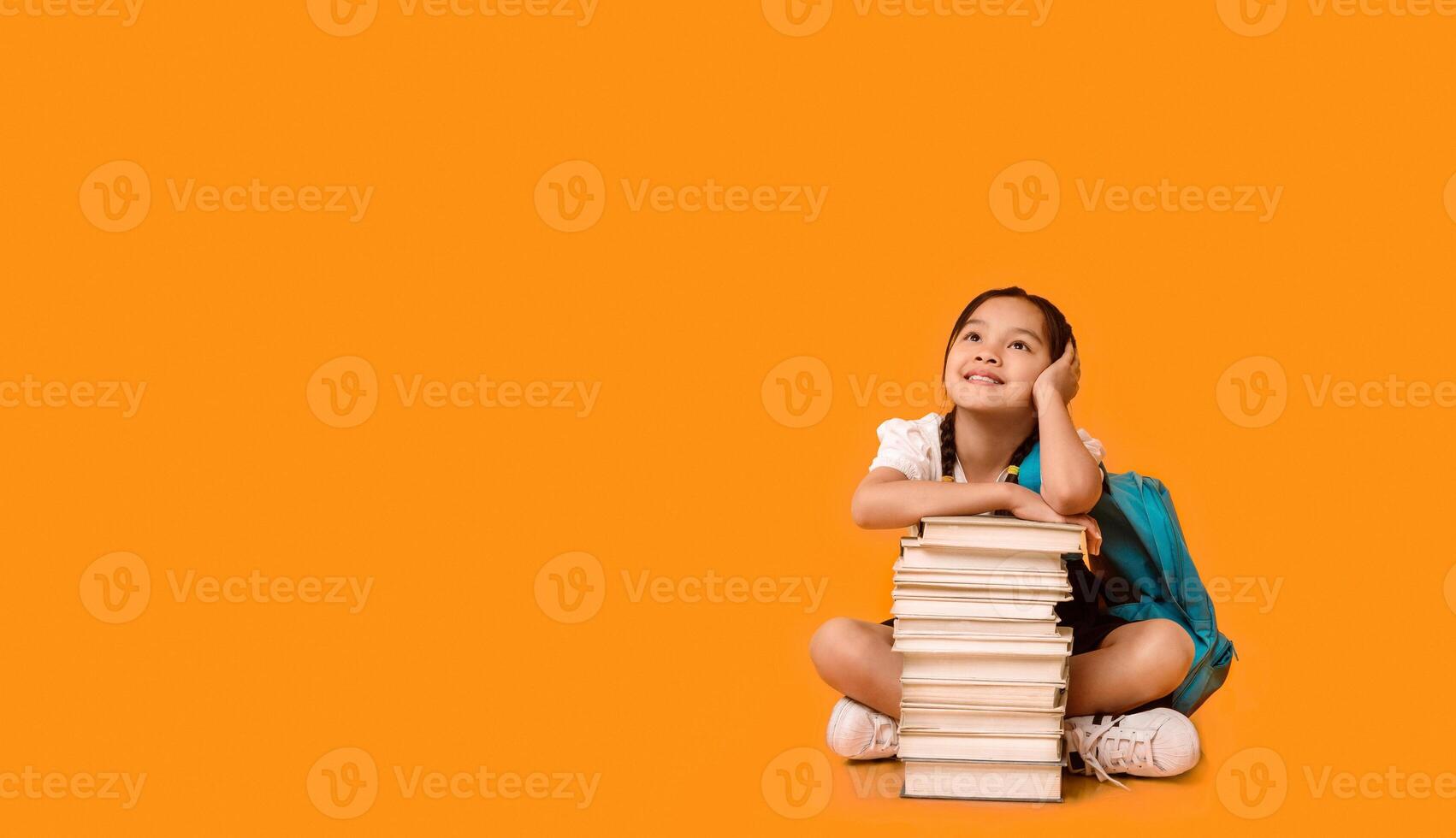Chinese Elementary School Girl Sitting At Books Stack Posing On Yellow Background. Studio Shot, Free Space For Text photo