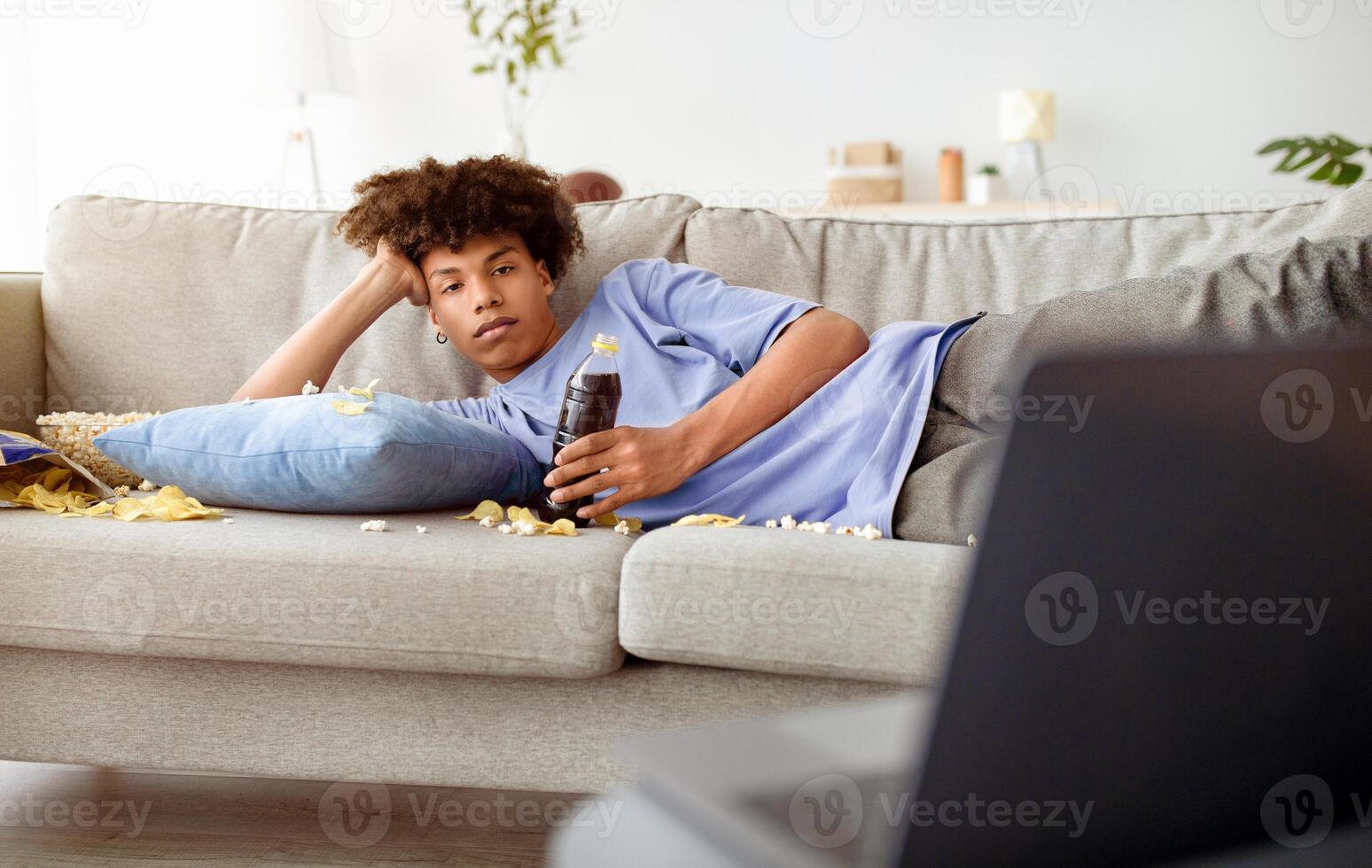 Bored teen guy lying on sofa with scattered snacks, watching movie on laptop at home. African American youth being lazy, eating popcorn and chips, enjoying interesting film online photo