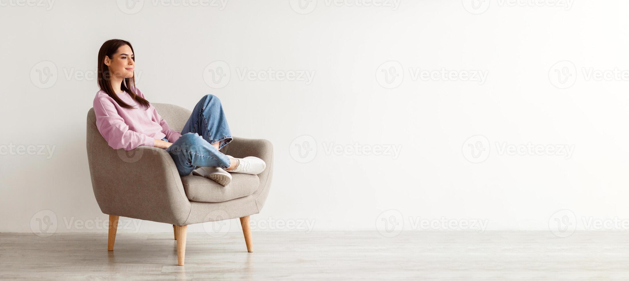 Young Caucasian woman relaxing in cozy armchair against white studio wall, copy space. Peaceful millennial lady having relaxing day, chilling on lazy morning, taking break from work photo