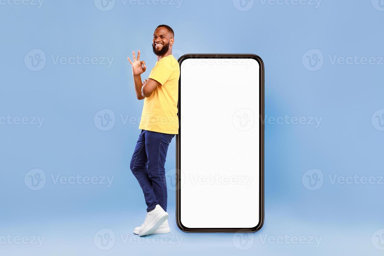 Optimistic African American man showing ok gesture, leaning on huge phone with blank white screen, recommending new mobile application on blue studio background, mockup for website ad design photo