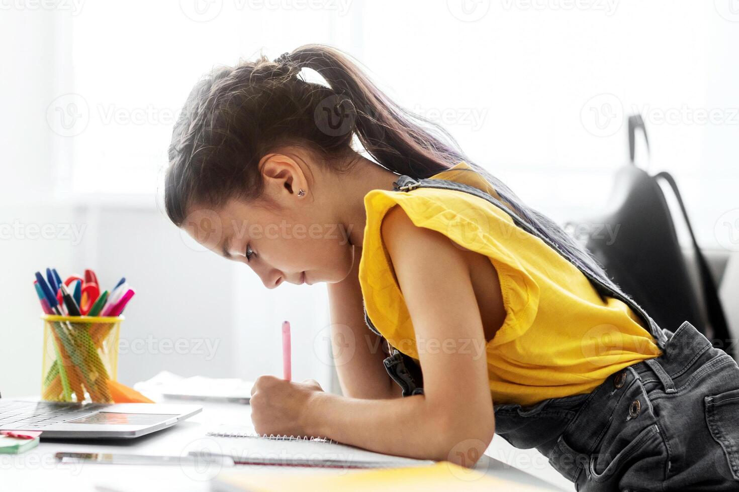 Distant Studying. Side view profile of focused little student writing down information watching webinar on laptop with empty screen, free space mock up. Youth making home assignment for online class photo