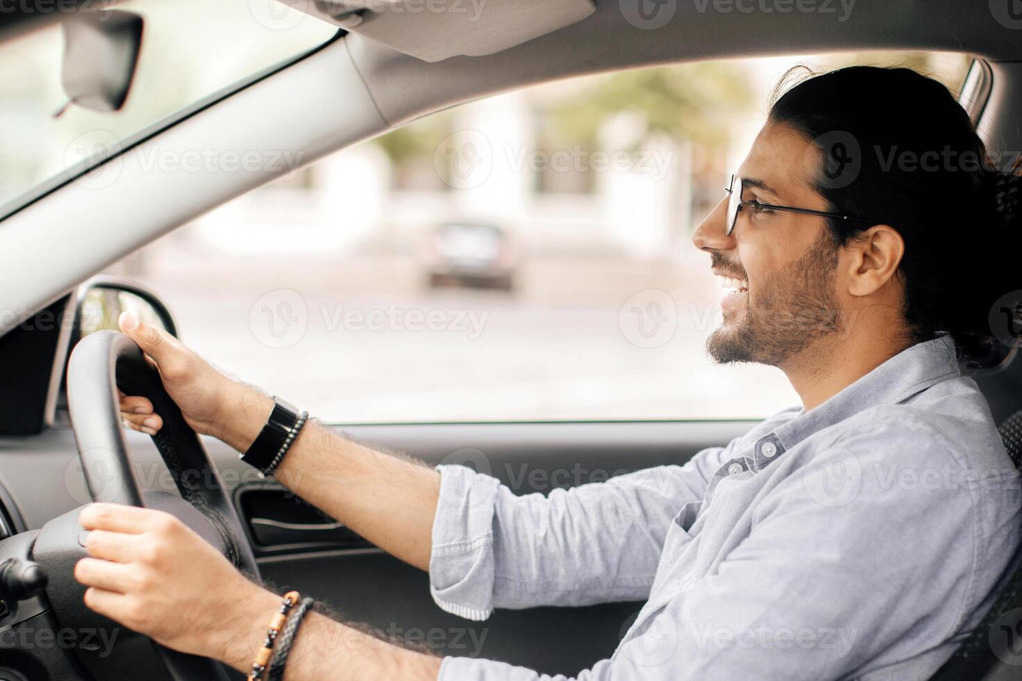 Handsome long-haired indian guy with glasses driving brand new car, looking at road and smiling, side view, copy space. Cheerful middle-eastern young man enjoying his new automobile photo