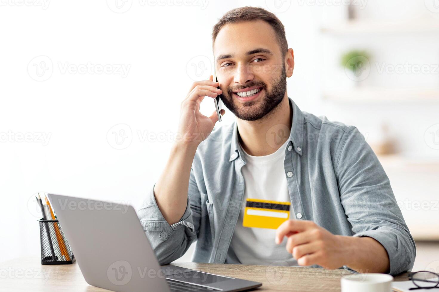 Cheerful young man buying things online, making phone call, using laptop and credit card for shopping in internet at home. Happy millennial guy purchasing goods on web, calling remote bank photo