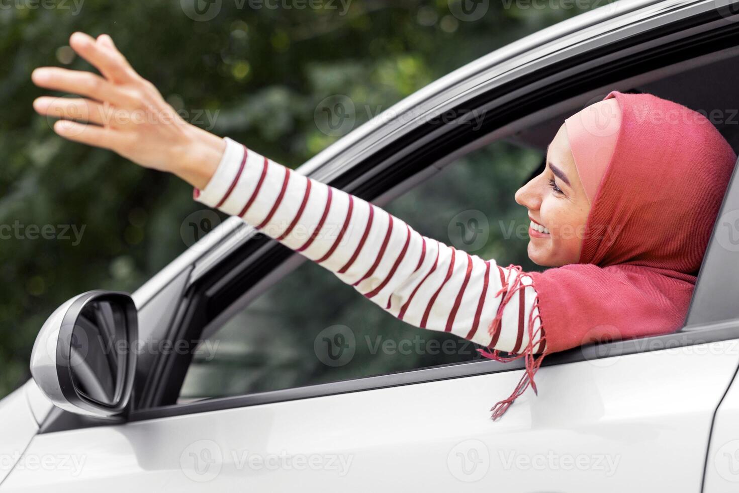 Satisfied millennial islamic woman in hijab catches air with her hand from open window in car, outdoor, panorama. Travel together, rest and trip on vacation by auto, good mood and freedom due covid-19 photo