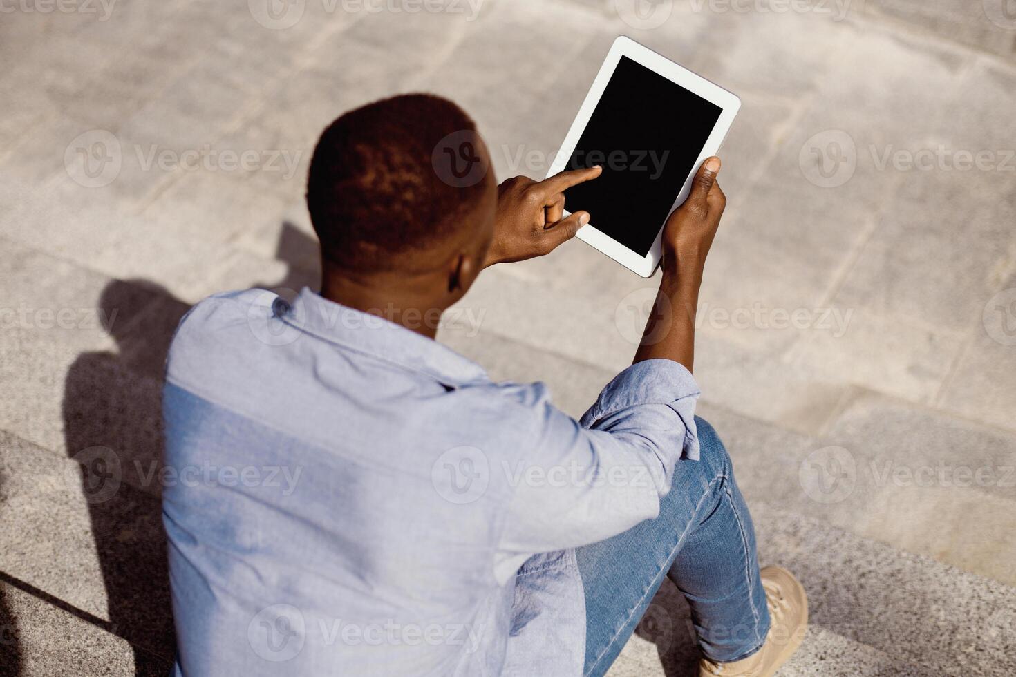 Young black guy using tablet computer with empty screen on stone stairs outdoors, mockup for website design. Millennial African American man pointing at touch pad with space for your template photo