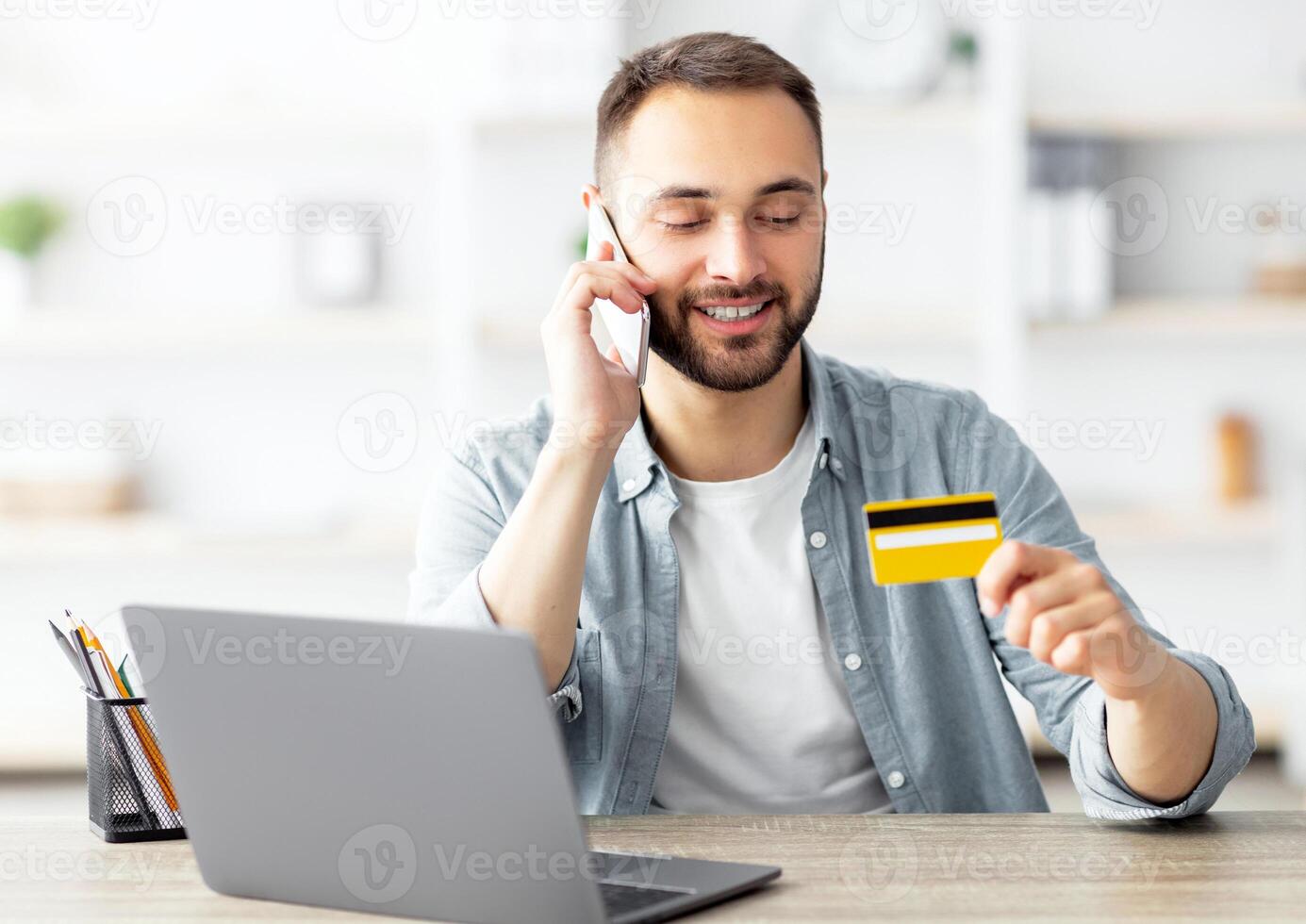 Cool young guy with laptop and credit card shopping online, making mobile phone call, confirming web order at home. Millennial Caucasian man making purchase over internet photo
