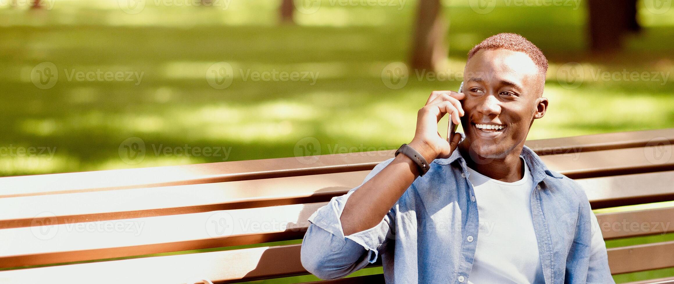 E-learning concept. Funky black student with laptop talking on smartphone on bench at park. African American guy talking to his college tutor while watching educational on pc, outside photo