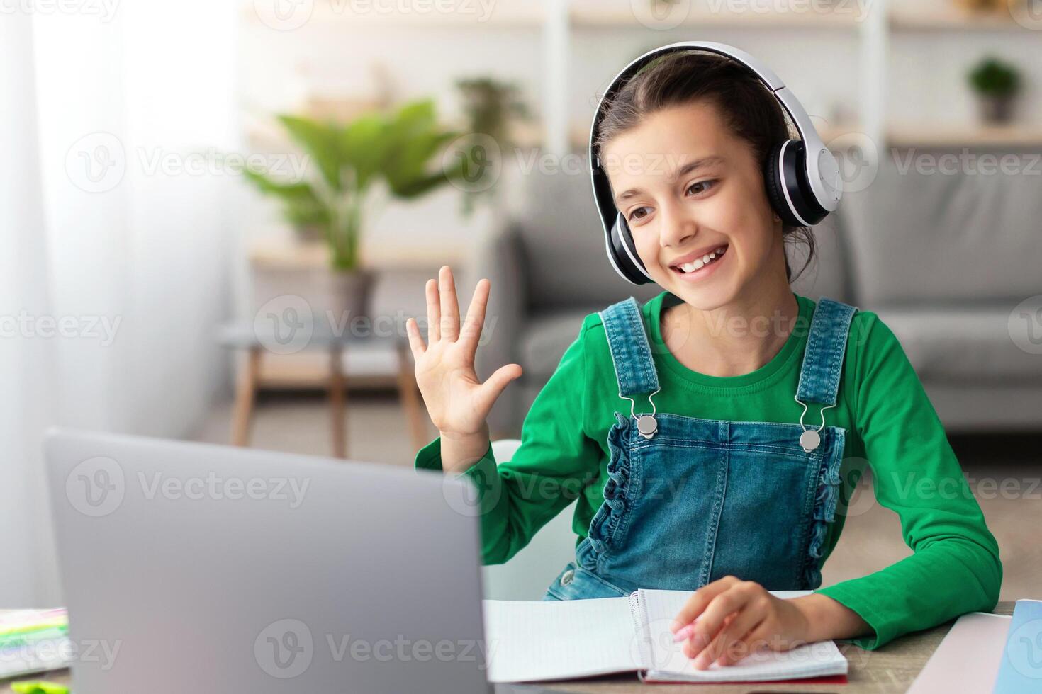 Distance Learning. Cute teenage girl in headset having lesson using laptop in living room, studying remotely, enjoying online education, waving hand at pc web camera photo
