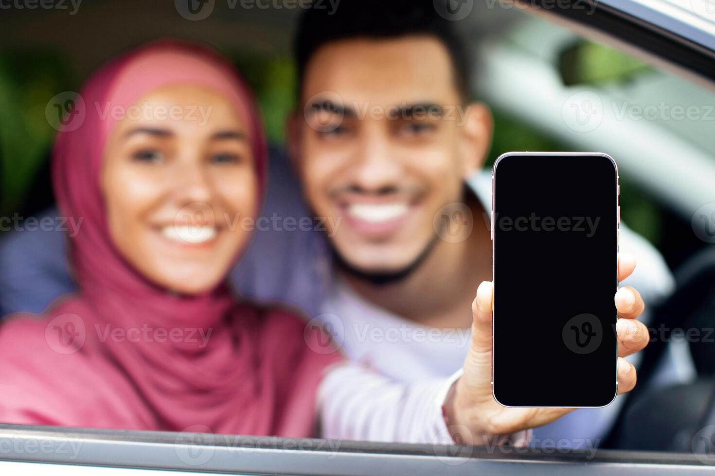 Online Car Insurance. Cheerful Muslim Couple Sitting In Auto And Demonstrating Blank Smartphone With White Screen, Happy Arab Husband And Wife In Hijab Recommending New Mobile Application, Mockup photo