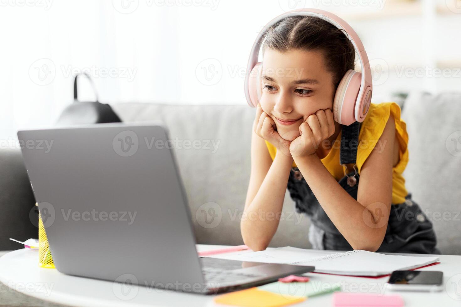 Kids And Technology Concept. Portrait of happy smiling teen girl using laptop computer, wearing wireless headphones, studying or watching movie online sitting on the couch in living room photo