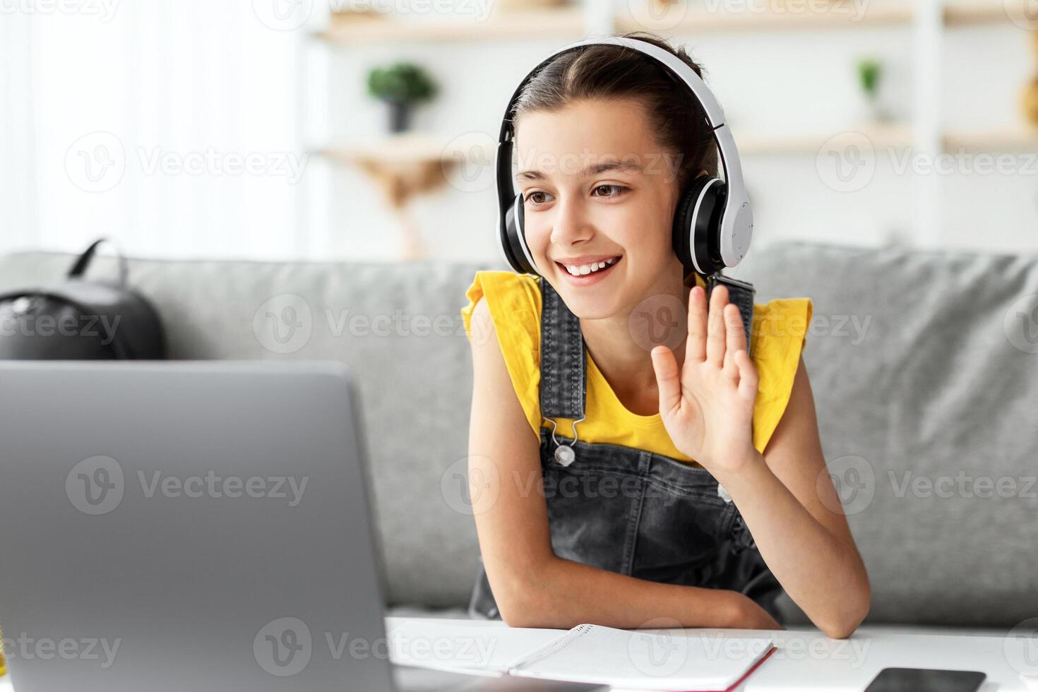 Remote Learning Concept. Portrait of positive girl in headset sitting at desk on couch in living room, using computer having virtual chat, waving hand to webcam greeting teacher and classmates photo