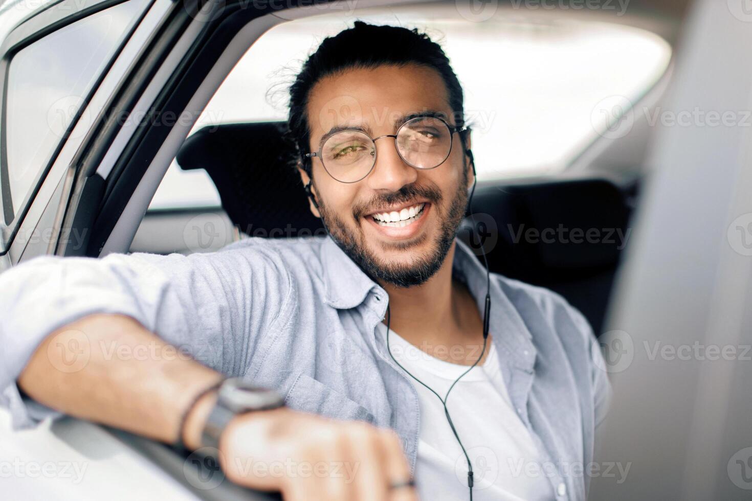 Stylish arab guy with glasses travelling by car service or taking taxi, using earphones, listening to music, enjoying auto ride on a back seat, looking through window and smiling, closeup photo
