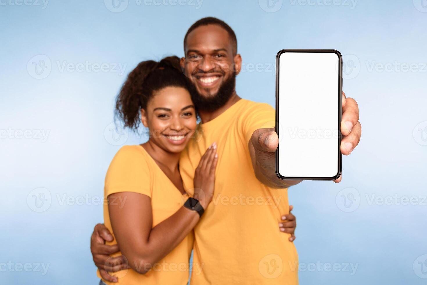 New awesome mobile app. African American couple presenting smartphone with mockup, promoting application or website, advertising your product or service, blue studio background. Selective focus photo
