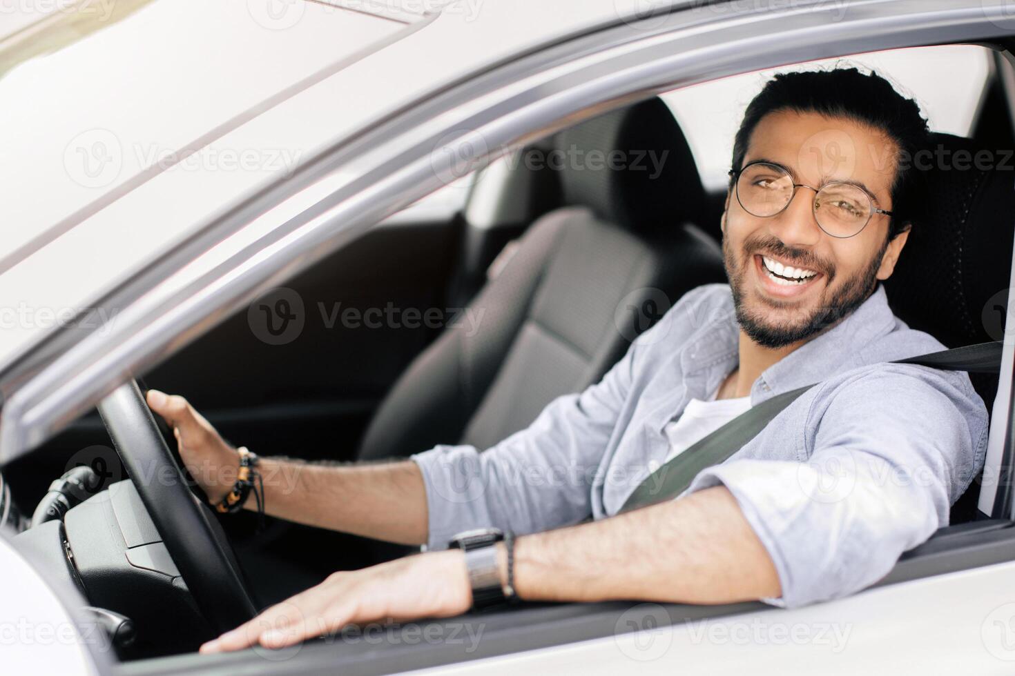 Cheerful long-haired arab guy with glasses driving right wheel auto, looking through open window and smiling at camera, happy young middle-eastern man enjoying car ride, copy space photo