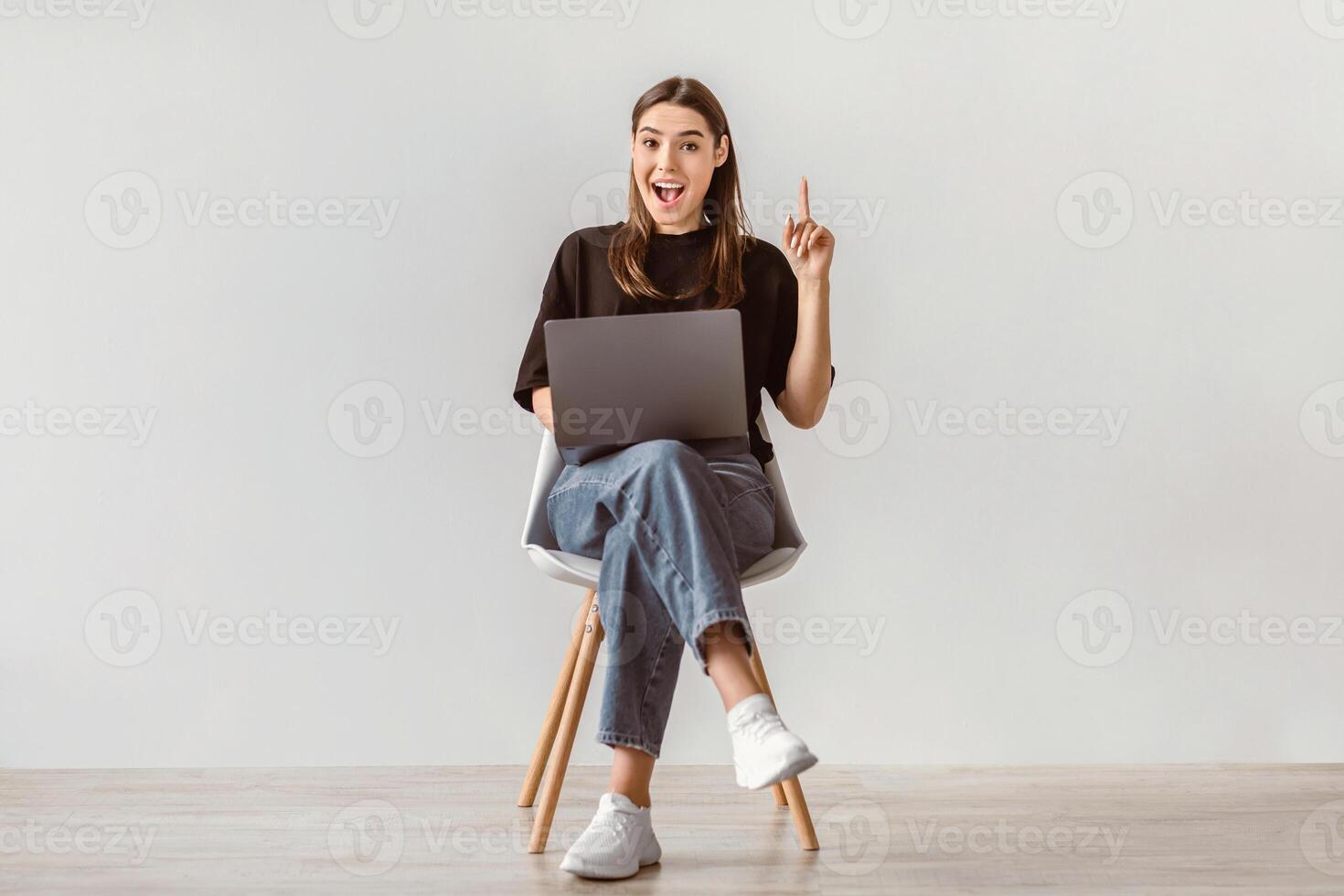 Full length of young Caucasian lady with laptop sitting on chair, pointing upwards, showing IDEA gesture against white wall. Millennial woman having aha moment, feeling excited during online work photo