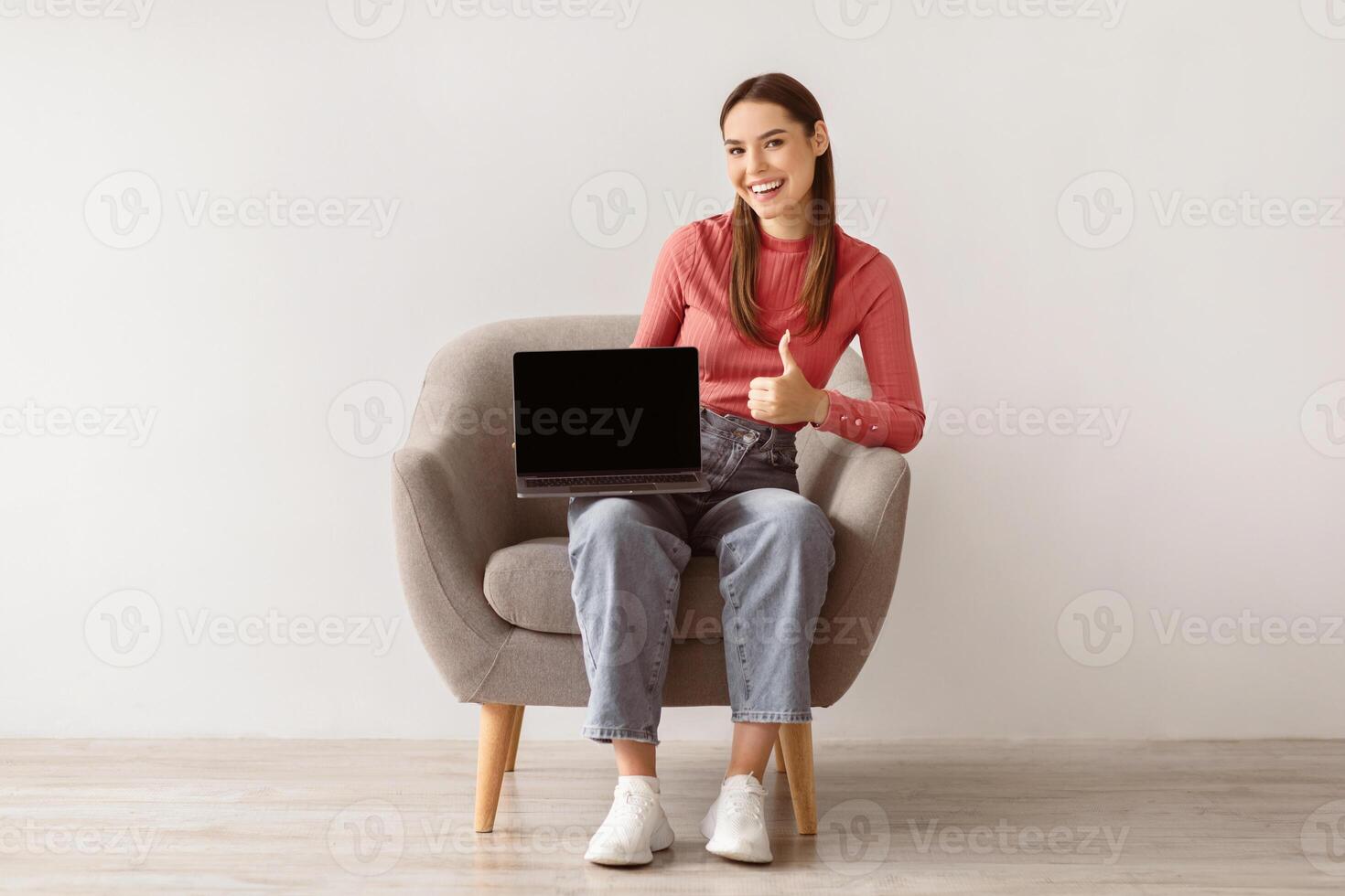 Happy young woman sitting in armchair, holding laptop with empty screen and showing thumb up, mockup for website. Positive Caucasian lady with portable computer, space for ad. Online work, education photo