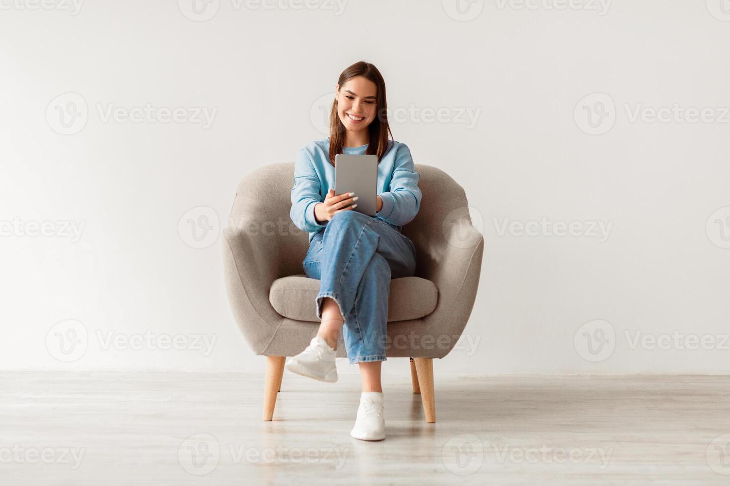 People, technology and communication concept. Portrait of smiling young woman with digital tablet sitting in armchair, working online against white studio wall, full length photo