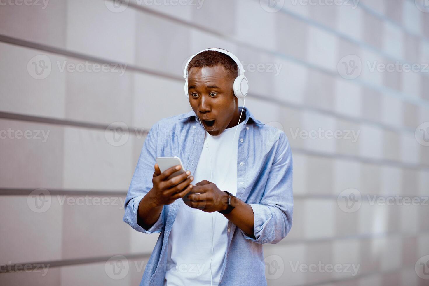 Shocked African American man in headset staring at smartphone screen on city street. Funky black guy surprised over huge sale or discount, unexpected news outdoors photo