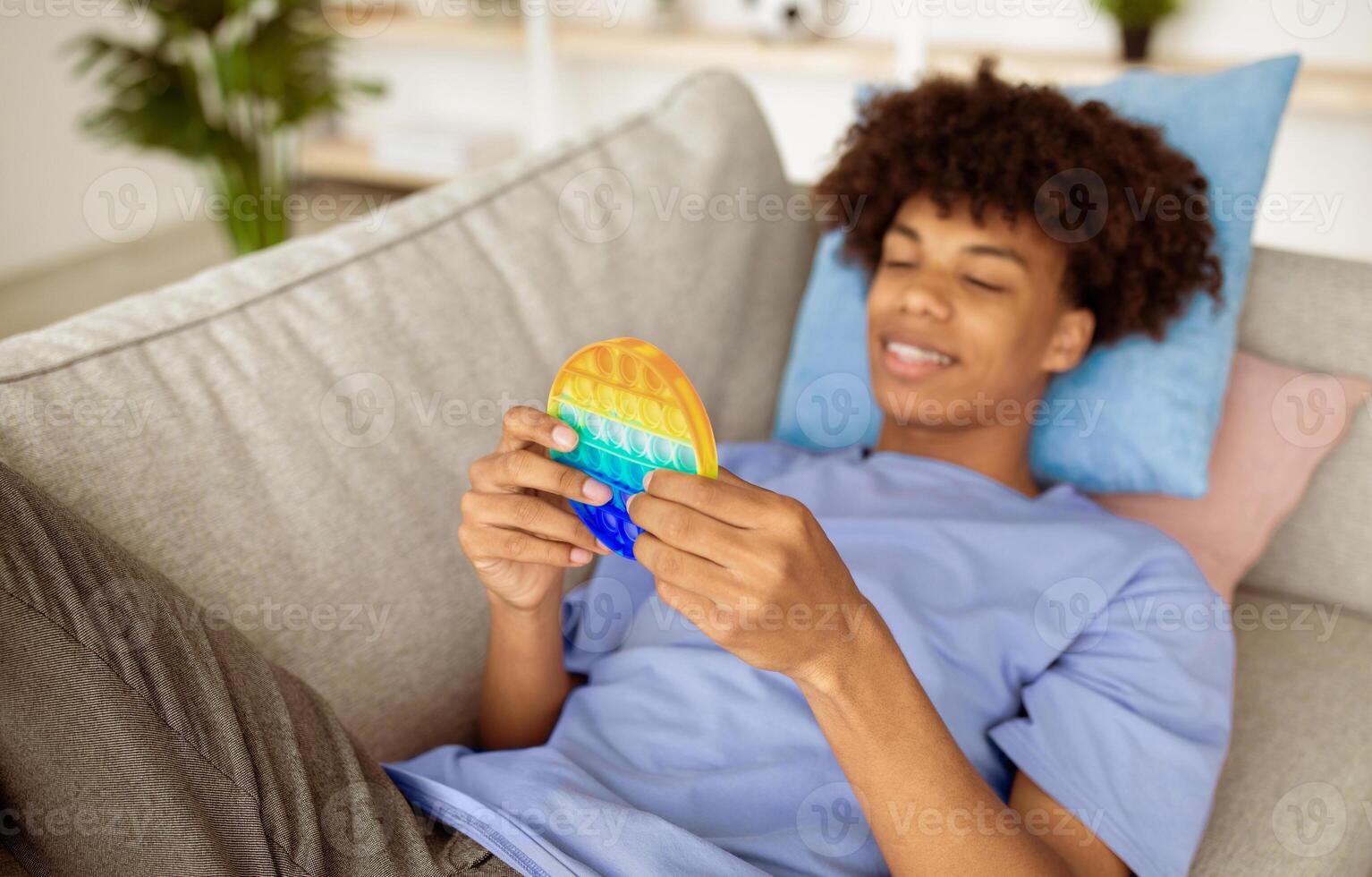 Cool black teen guy playing with bright POP IT toy, enjoying fun antistress game at home, selective focus. African American adolescent trying out modern popular plaything photo