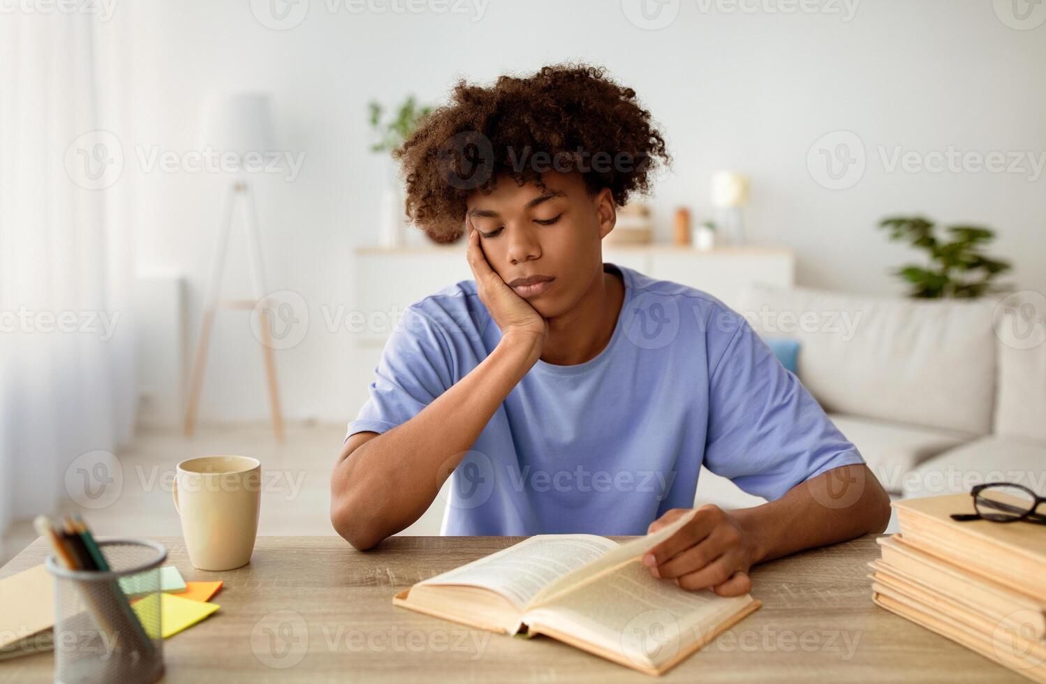 Bored black teenager sitting at desk, reading book, studying at home. African American youth learning remotely, making assignment, using textbook to prepare for exam, indoors photo