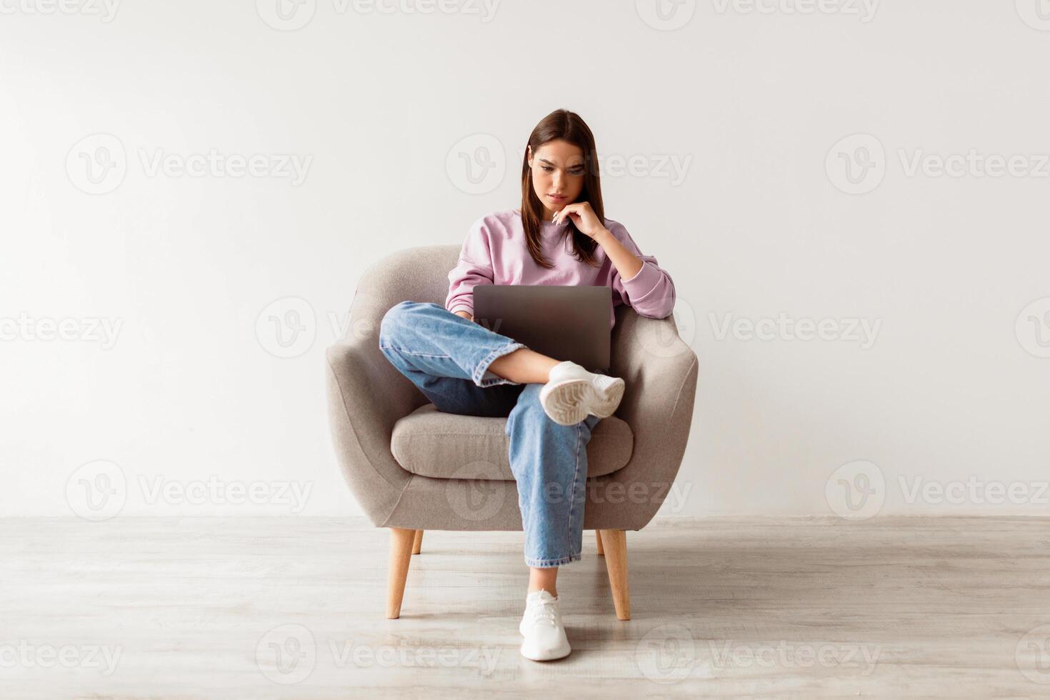 Serious young lady sitting in armchair with laptop computer, studying remotely or conferencing against white studio wall, full length. Millennial woman watching webinar, having online job photo