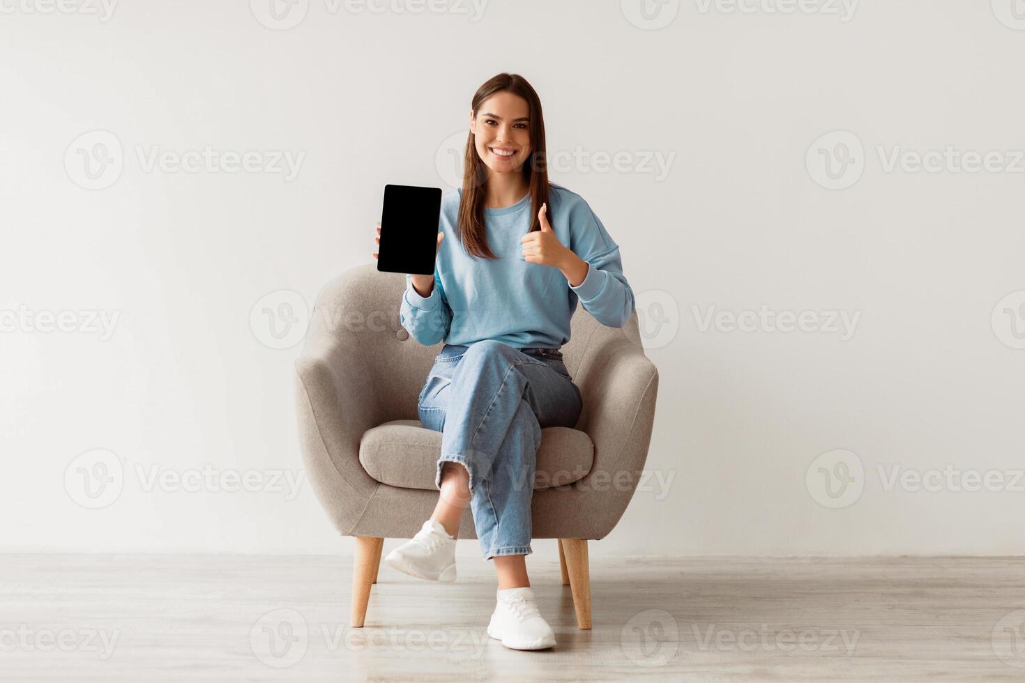 Smiling millennial woman showing empty touch pad, gesturing thumb up, sitting in armchair against white wall, mockup for website design on tablet screen. Online ad concept photo