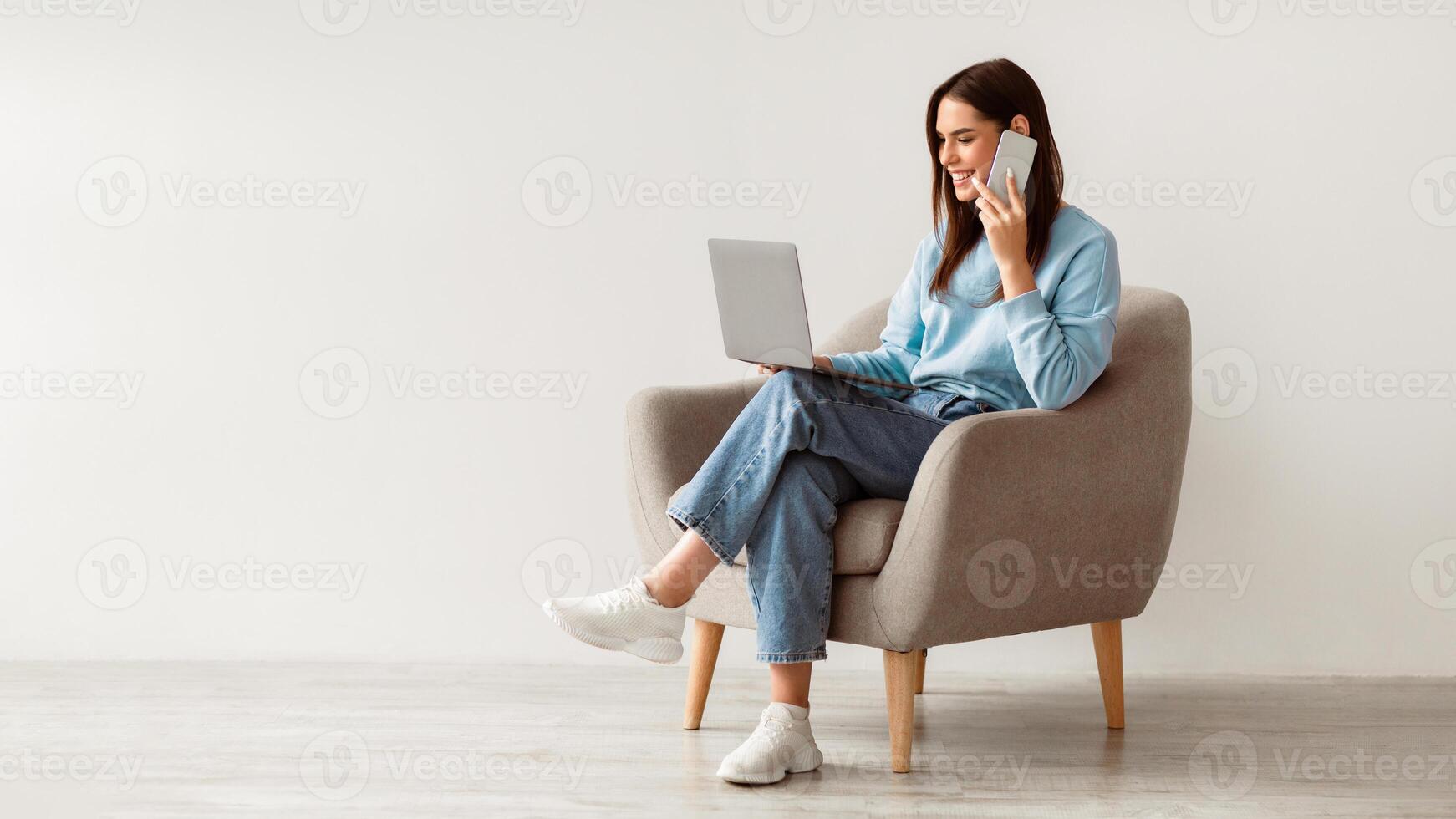 Full length of Caucasian lady using laptop and speaking on smartphone while sitting in armchair against white studio wall, free space. Lovely young woman having phone conversation, working on pc photo