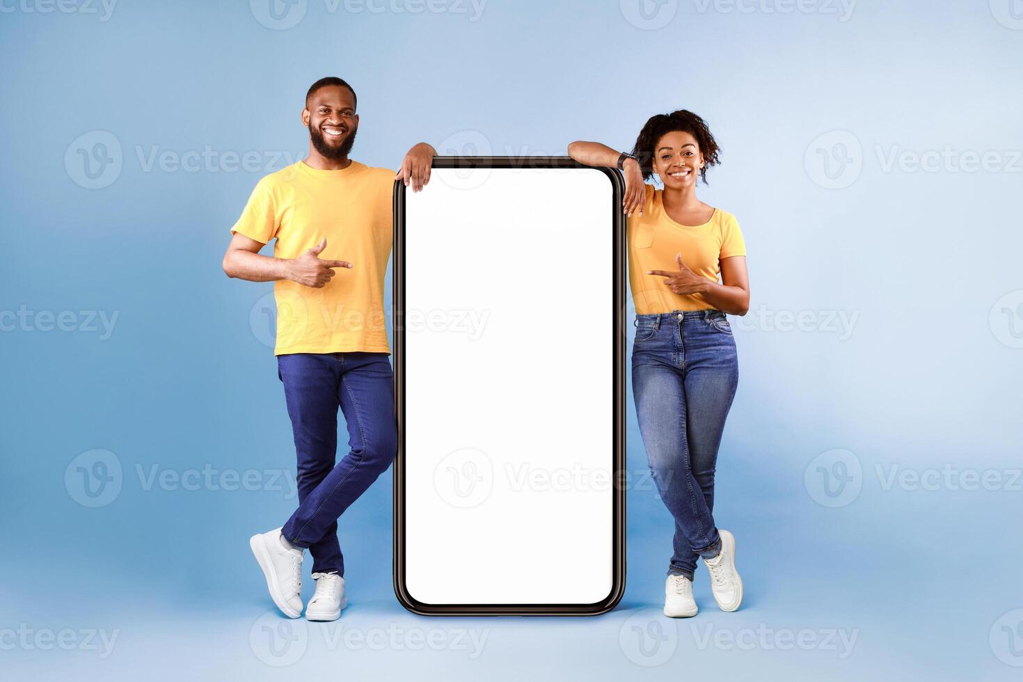 Great mobile app. African American couple pointing at giant smartphone with mockup, promoting application or website, advertising product or service, blue background. Full length photo