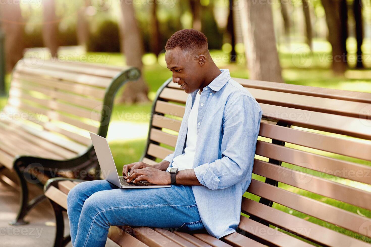 Portrait of cool black guy using laptop on bench at park, working remotely, communicating on webcam. Funky young student participating in online class or web conference outdoors photo