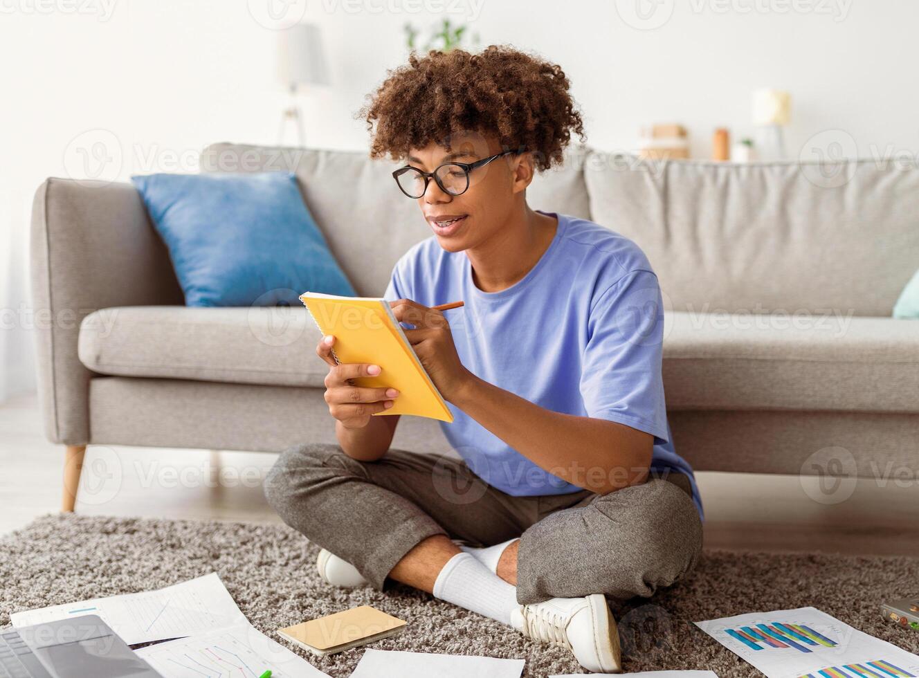 Full length of smart black teen student with laptop taking notes during online lesson at home. Intelligent teenage guy writing assignment or coursework paper, studying remotely indoors photo