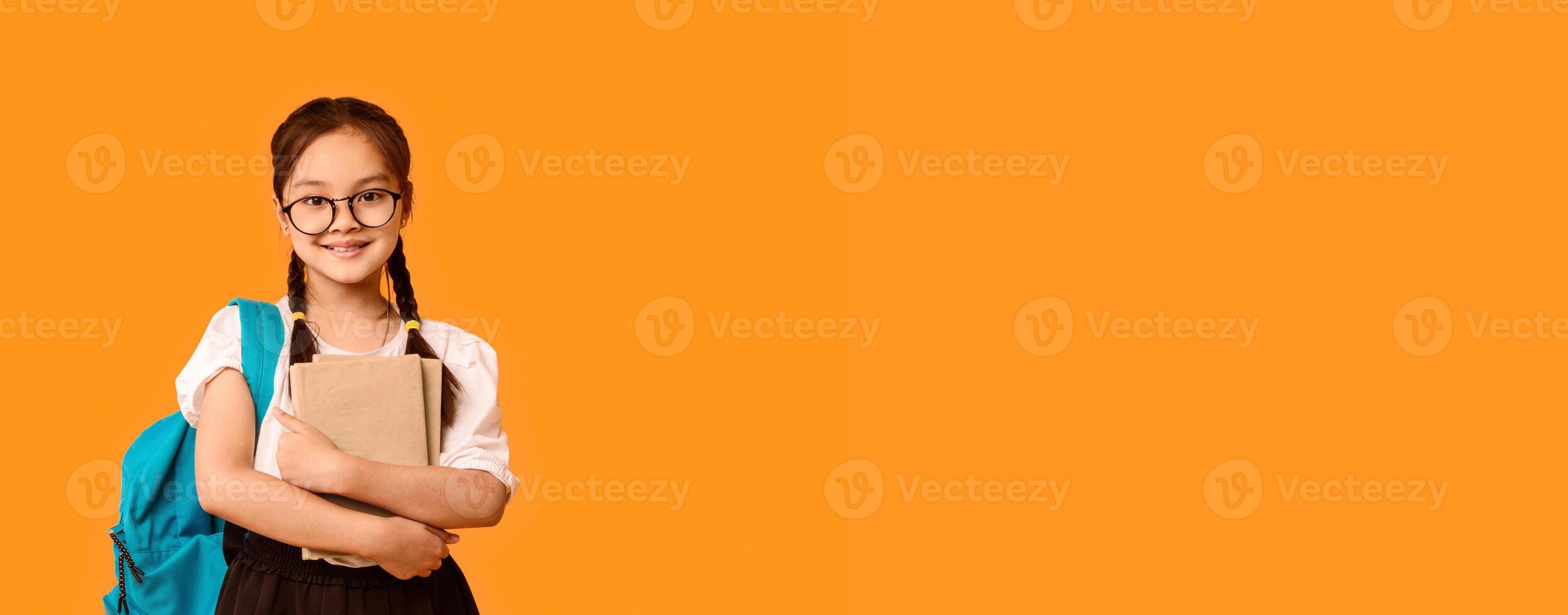 Back To School. Happy Asian Schoolgirl Embracing Book Standing With Backpack In Studio On Yellow Background. Free Space For Text photo