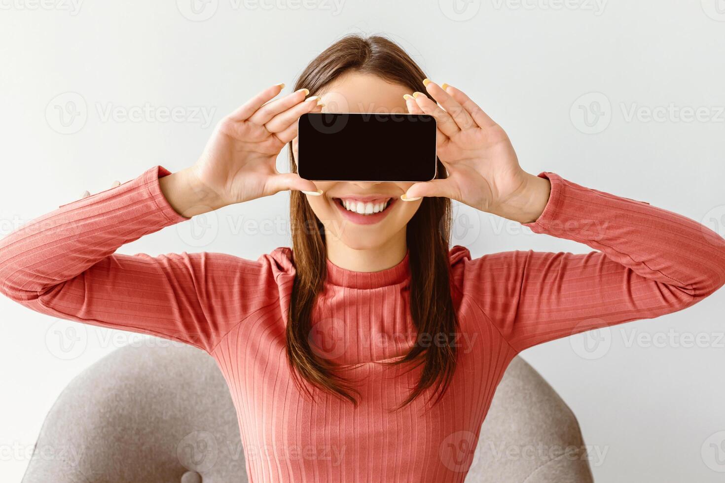 People and gadgets. Cheery young lady covering her eyes with cellphone on white studio background, mockup for app or website on gadget screen. Millennial woman offering space for ad on touch pad photo