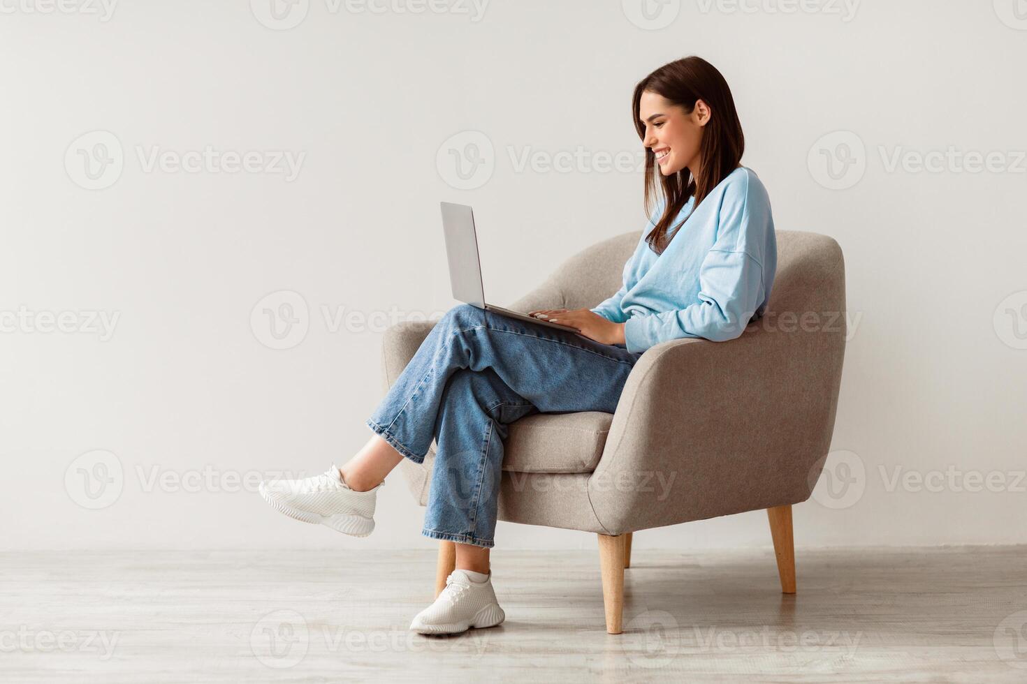 Smiling young woman in casual wear working online, sitting in armchair and using laptop against white studio wall, copy space. Cheerful Caucasian lady surfing internet on portable pc photo