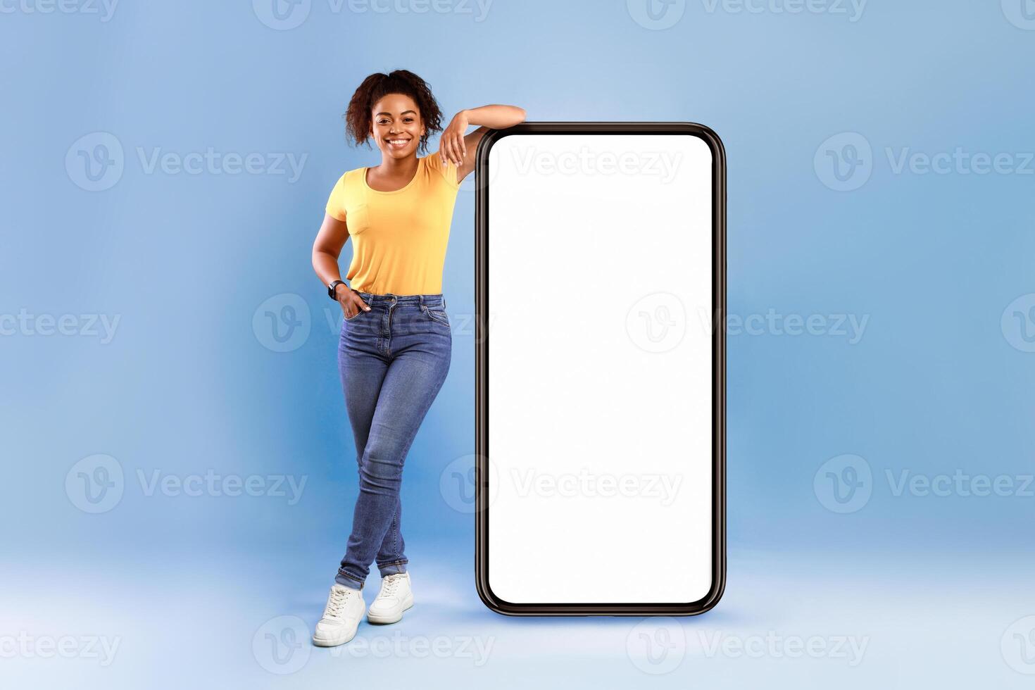 Mobile app advertisement. Happy African American woman leaning on huge smartphone with empty white screen, advertising new website or application, blue studio background photo