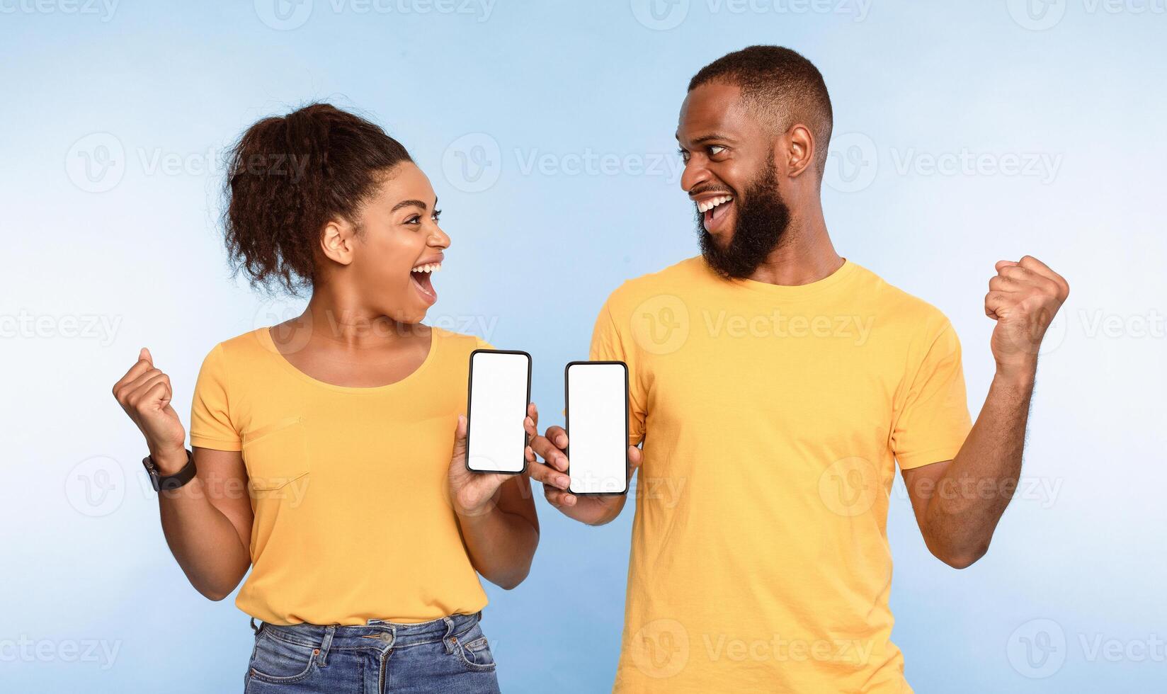 Excited over this new mobile app. Overjoyed African American couple holding smartphones with mockup for design, making YES gestures, advertising website over blue studio background photo