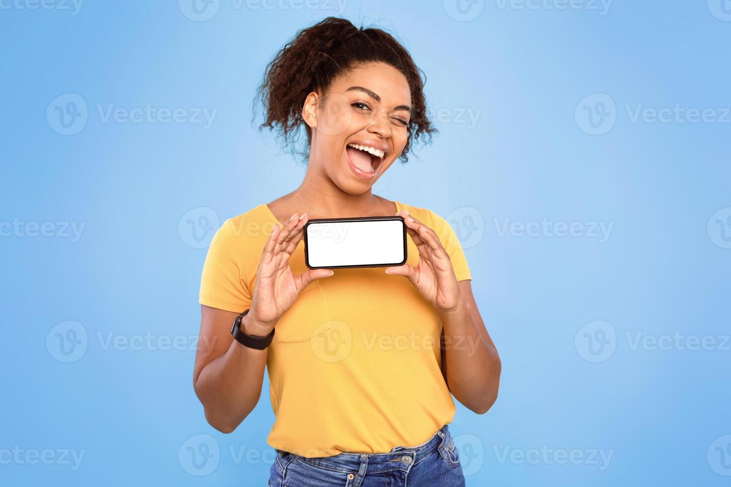 Pretty African American lady winking and holding smartphone with blank screen on blue background, mockup. Young woman offering new mobile app, advertising website, copy space photo