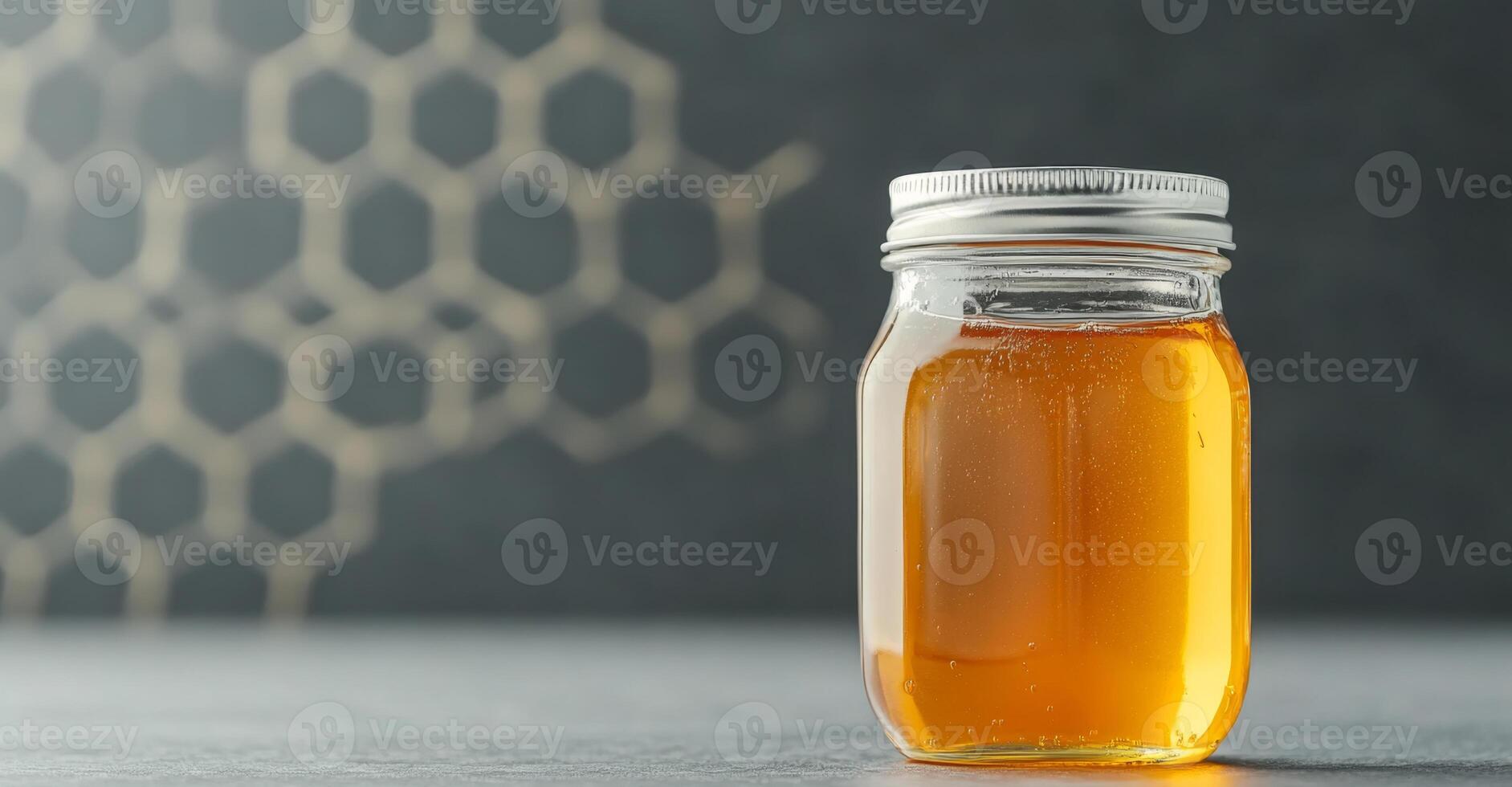A clear jar of honey sits on a table with a sleek design, surrounded by a subtle honeycomb pattern in the background. This organic treat highlights the beauty of nature's sweetness. photo