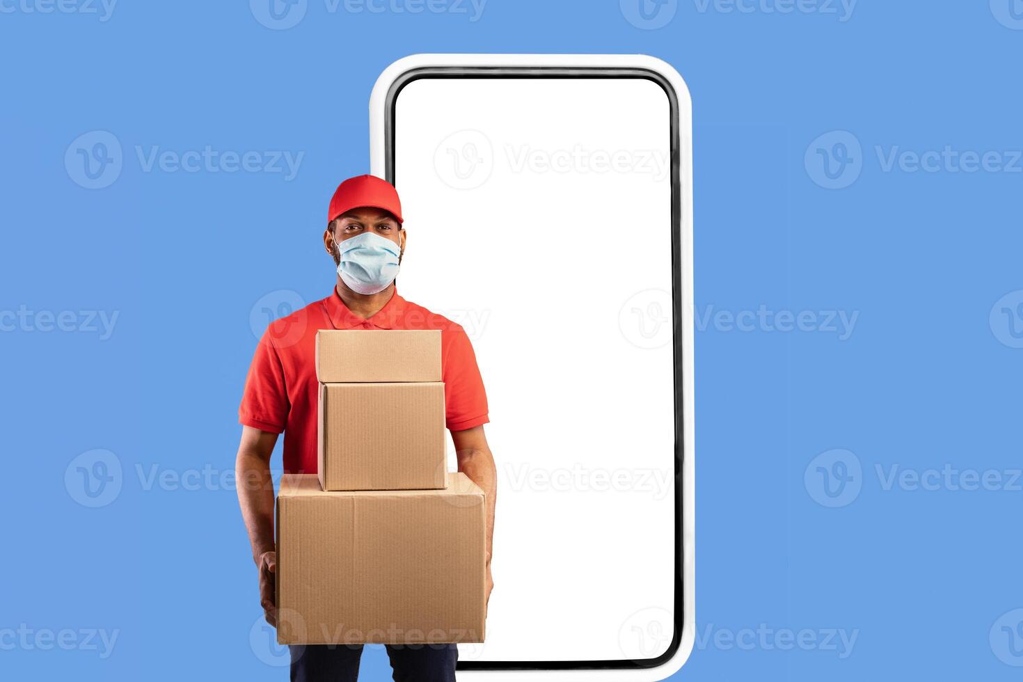 Delivery Application. African American Courier Standing Near Big Phone Screen Holding Cardboard Boxes Looking At Camera Over Yellow Studio Background, Wearing Face Mask. Square photo