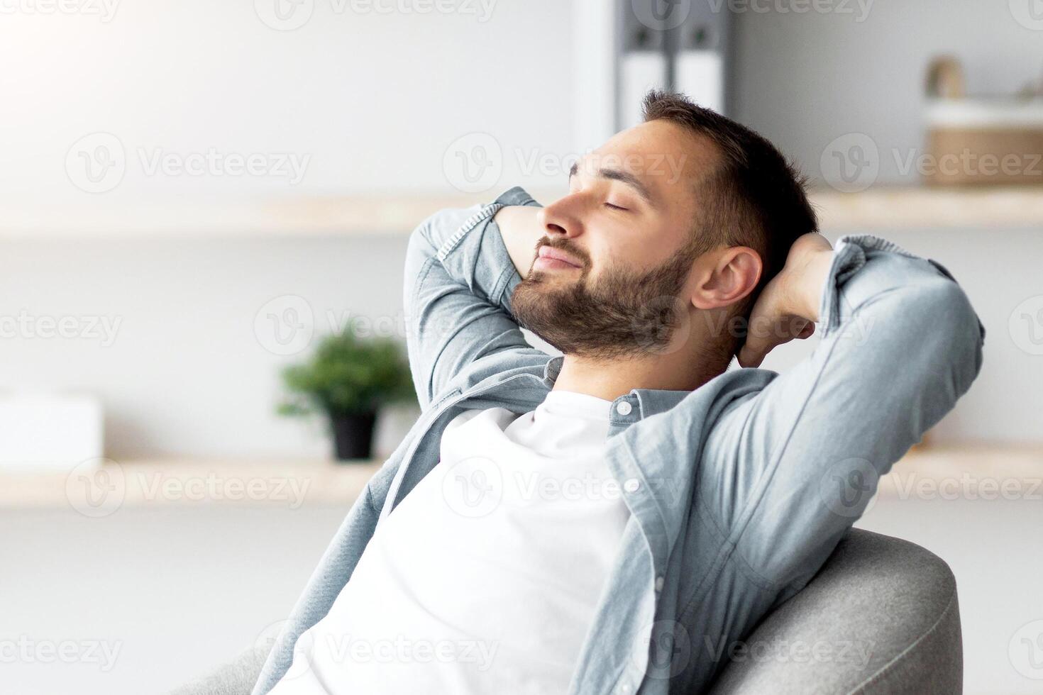 Cheerful young man resting in comfy armchair, chilling with closed eyes, holding hands behind his head at home. Positive Caucasian guy spending relaxing weekend morning in living room photo