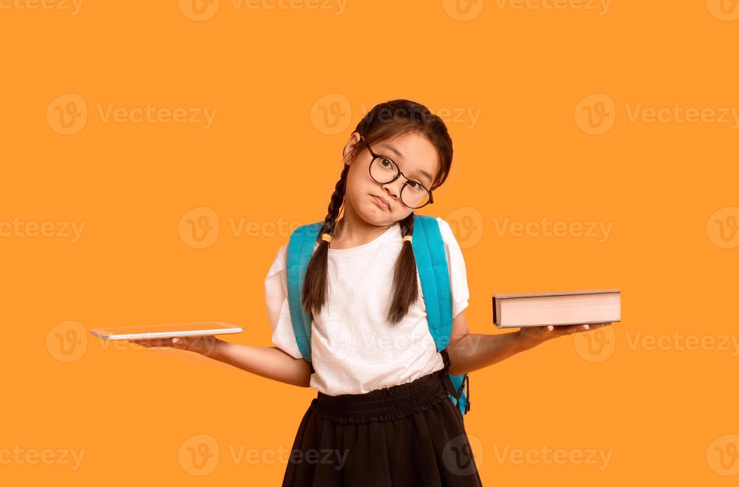Puzzled Chinese Schoolgirl Comparing Digital Tablet And Book Posing In Studio Over Yellow Background. Books Vs Gadgets Concept photo