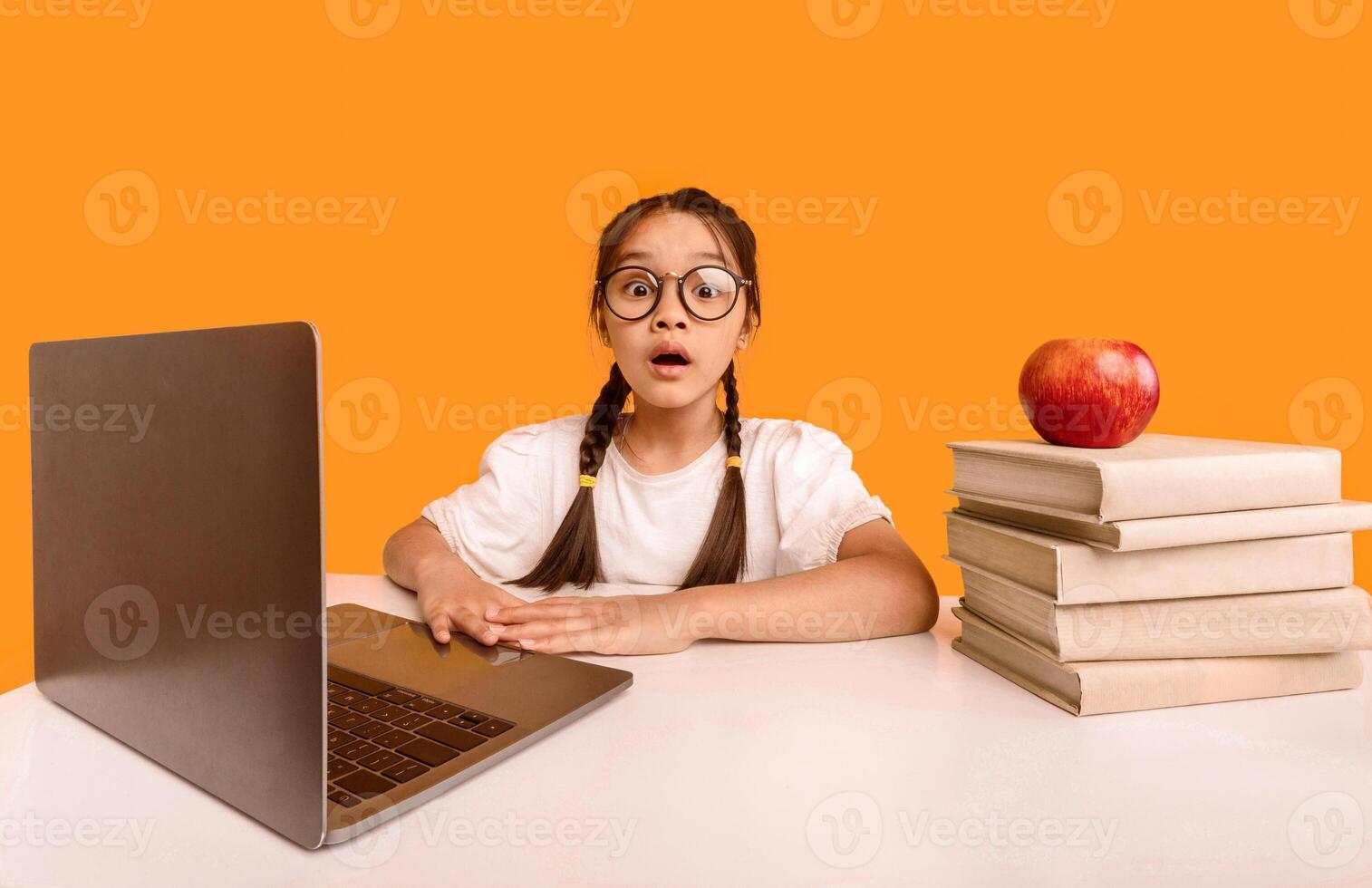 Shocked Japanese Elementary School Girl At Laptop Looking At Camera Sitting At Desk During Online Class Over Yellow Studio Background. photo