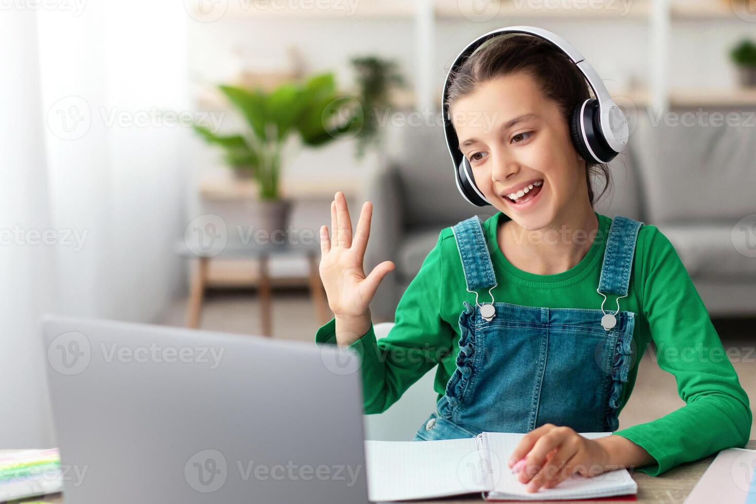 Online Call And Communication. Portrait of cheerful teenage pupil sitting at table, using laptop having virtual conference, waving hand to webcam greeting teacher and classmates, writing photo